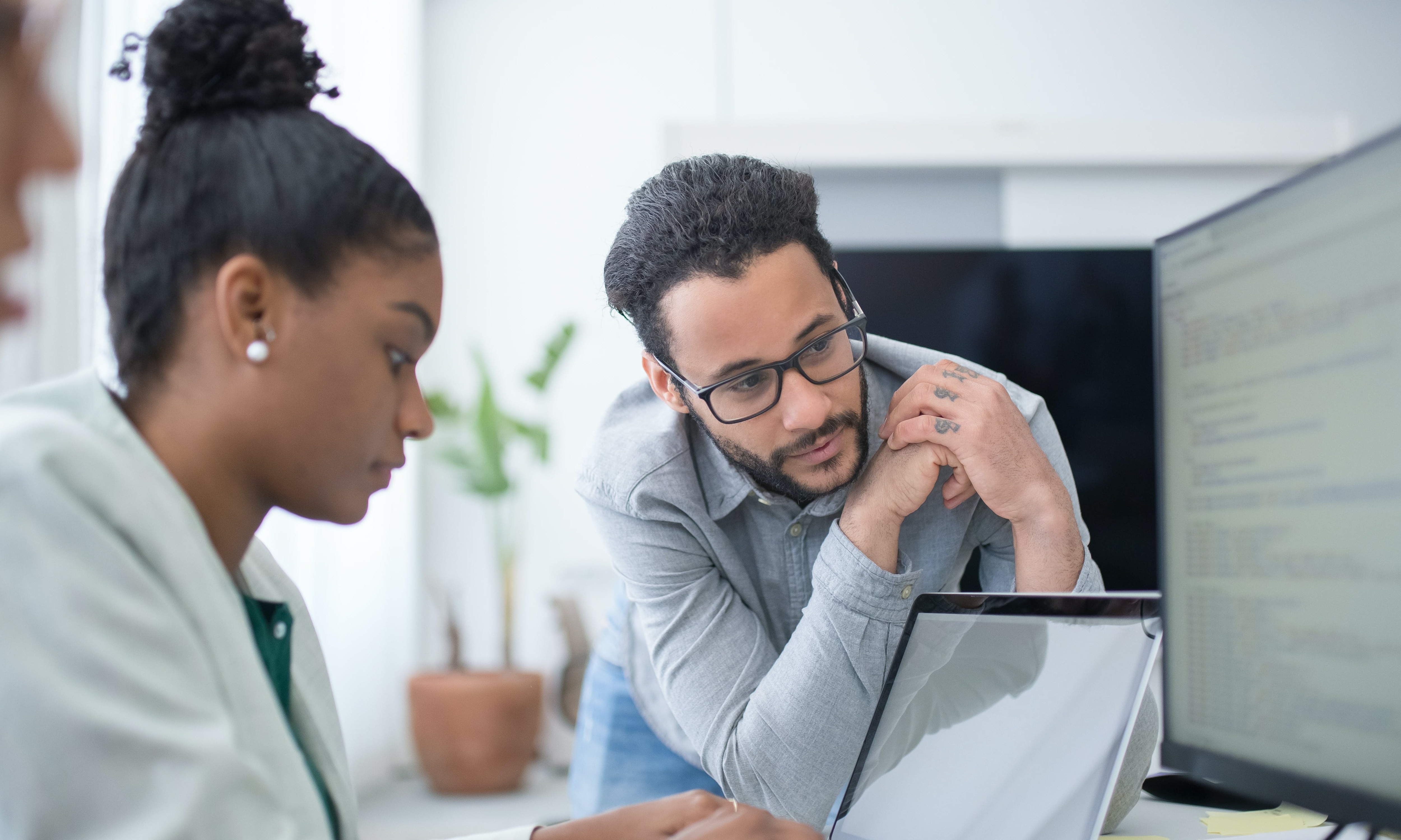 Two office workers, a woman looking at a laptop, and a man watching over, looking at the bigger monitor. 