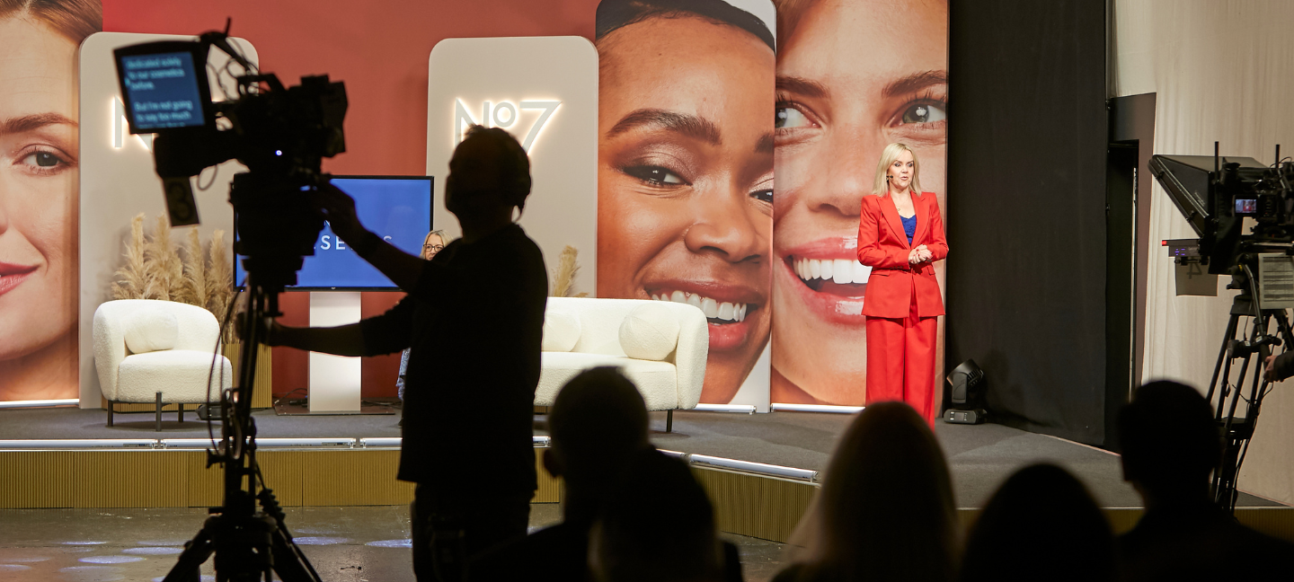 BTS shot of a camera filming a female presenter dressed in red at the No 7 Virtual Training Event