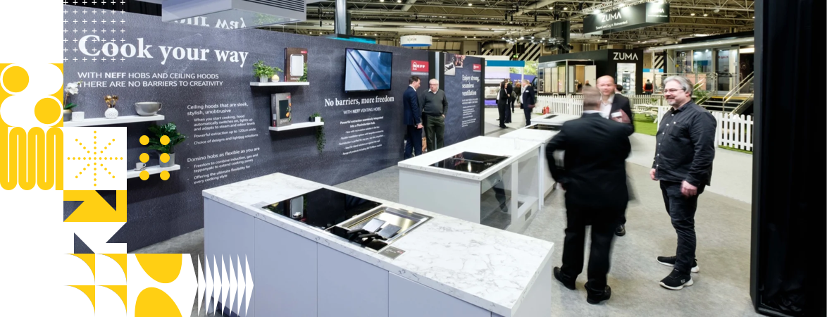 Exhibition space for NEFF kitchen appliances. 5 people on a stand with three islands with cooking and a wall with TV screen and four shelfs with items 