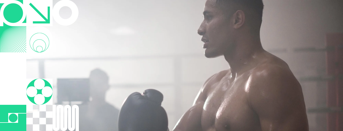 English Boxer Delicious Orie wearing boxing gloves in training. In the background is a blured image of a camera and producer. Left hand side is mix of white and green boxes with DRPG branding