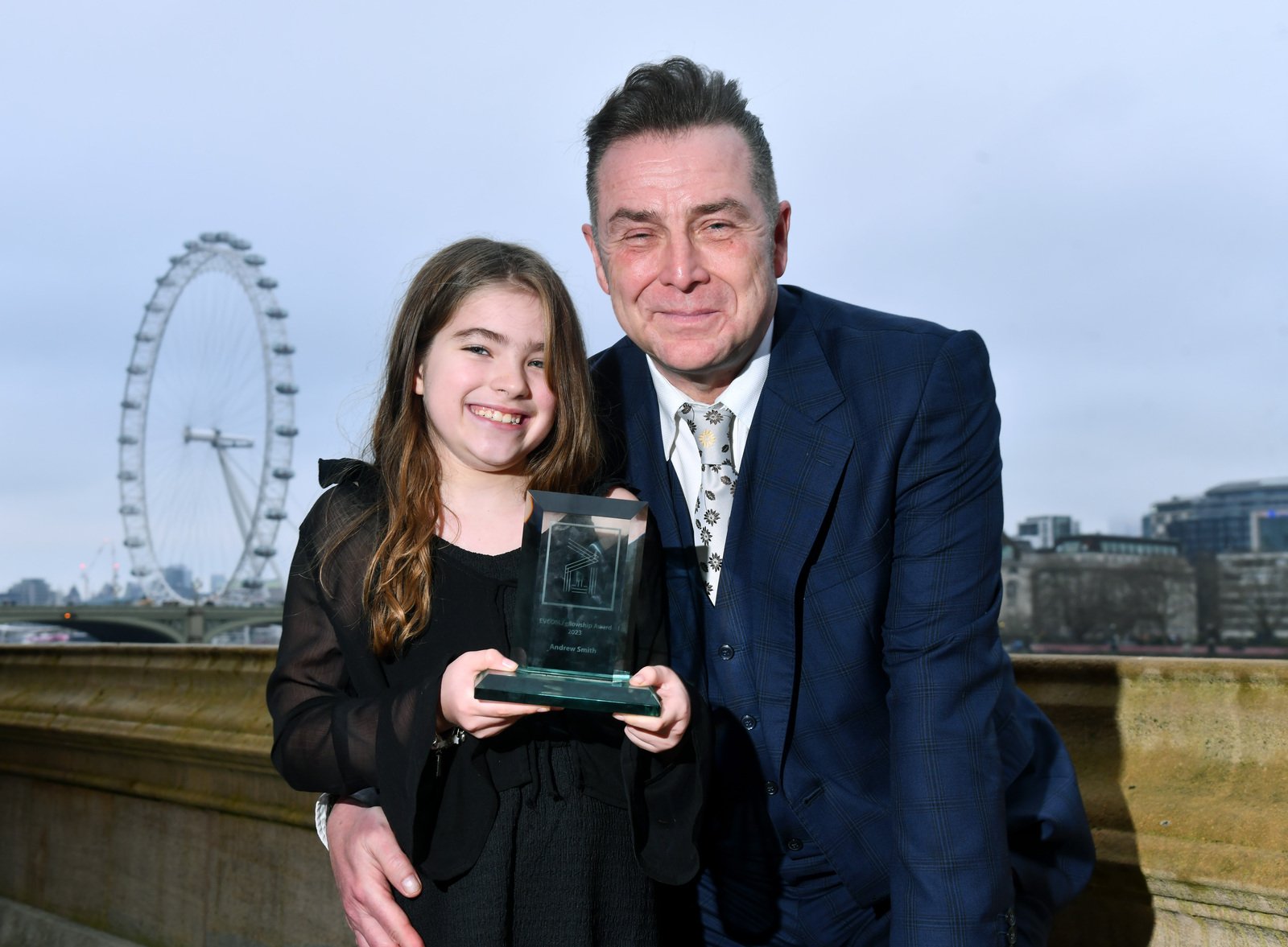 Andrew Smith stands alongside his daughter who is holding the trophy in front of the London eye at the EVCOM Fellowship Awards.
